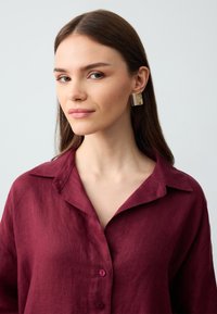 Bordowa lniana koszula z kołnierzykiem i guzikami; model nosi prostokątne kolczyki z przezroczystym wzorem i kolorowymi paskami.