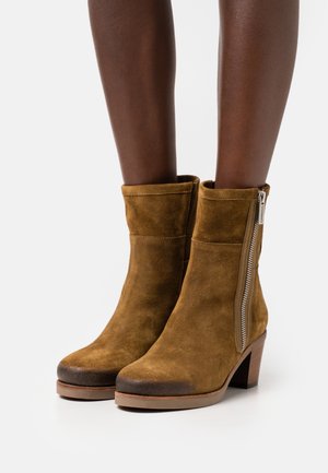 Botas de ante marrón hasta el tobillo con cremallera lateral, punta redonda y tacón acampanado. Textura suave con un acabado liso.