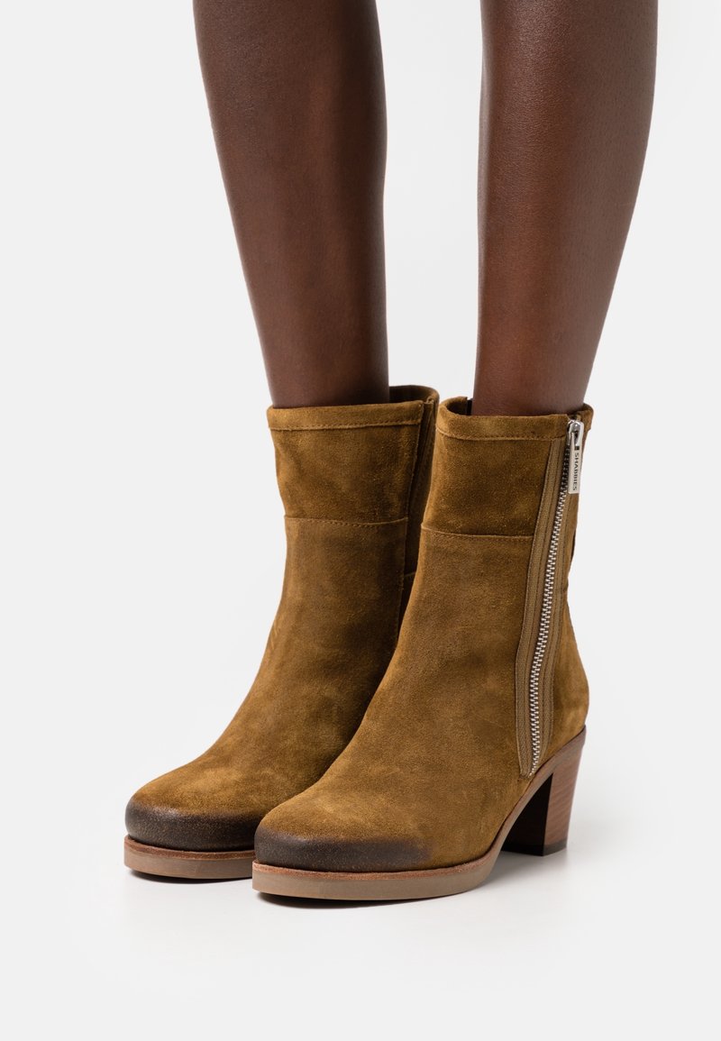 Botas de ante marrón hasta el tobillo con cremallera lateral, punta redonda y tacón acampanado. Textura suave con un acabado liso.