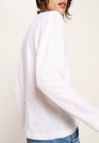 Frau, die ein locker sitzendes weißes Langarmshirt und blaue Jeans trägt, seitlich vor einem hellen, einfarbigen Hintergrund gezeigt.