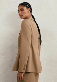 Smėlio spalvos prigludęs švarkelis su aptemptu siluetu, ryškiomis atlapomis ir keturių mygtukų detalėmis ant rankovės. Lygus audinys su struktūruotu siluetu.