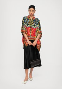 Multicolored patterned kaftan with short sleeves, featuring red, blue, and yellow designs, paired with a black pleated skirt and handbag.