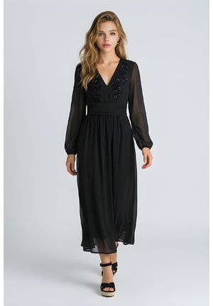 Joven mujer con cabello largo, vestida con un vestido negro largo con mangas transparentes y bordados florales que camina hacia adelante sobre un fondo liso.