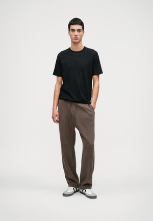 Černé tričko s kulatým výstřihem v kombinaci s hnědými tepláky s šňůrkou. Model má na sobě bílo-šedé tenisky.