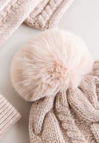 Beige knitted hat with a textured cable pattern and a fluffy pale pink pom-pom on top. Soft material with a cozy appearance.