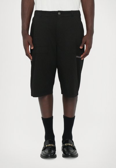 Shorts cargo noirs à coupe ample, dotés de deux poches latérales et d'un logo brodé sur la jambe. Portés avec des chaussettes noires et des mocassins brillants.