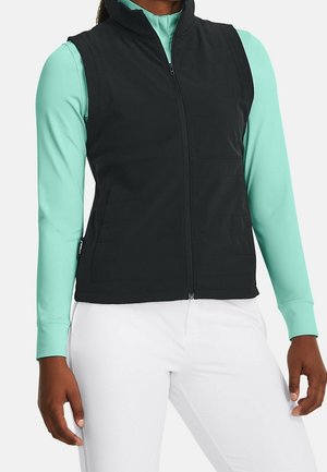 Persoon draagt een zwart vest met rits over een teal gekleurde top met lange mouwen en witte broek, staat met handen ontspannen langs het lichaam.