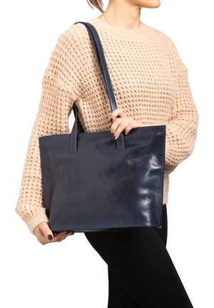Sac fourre-tout en cuir bleu marine avec deux poignées supérieures, forme rectangulaire, texture lisse et détails de couture minimalistes, tenu par une personne portant un pull.