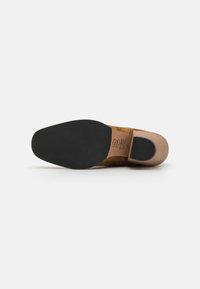 Semelle en caoutchouc lisse d'une chaussure en cuir suédé marron, avec un bout arrondi et un design plat, accentuée par un logo estampé sur la semelle intérieure.