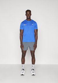 Zils sporta T-krekls ar īsām piedurknēm, ar logotipa detaļu uz krūtīm, kombinēts ar pelēkiem šortiem, baltiem sporta apaviem un pelēkām zeķēm.