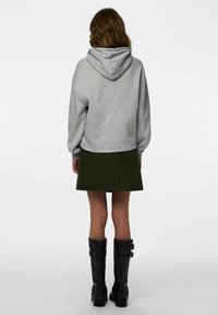 Pelēks sporta krekls ar kapuci, brīvs piegriezums, kopā ar tumši zaļu svārku. Melni ceļgalu augstuma zābaki ar sprādžu akcentiem. Minimālistisks fons.