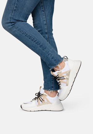 Baskets blanches et dorées avec des panneaux en mesh, des lacets noirs et une semelle épaisse, associées à un jean en denim bleu déchiré.