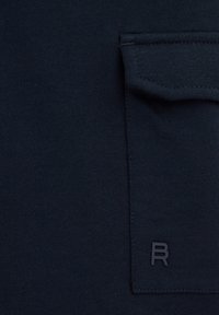 Tissu bleu marine avec une poche plaquée texturée, portant un petit logo en relief "R" dans le coin inférieur droit. Matériau doux et extensible.