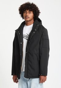 Jeune homme aux cheveux bouclés porte une veste noire à capuche par-dessus un T-shirt blanc à motif graphique et un jean bleu clair, debout devant un fond blanc uni.