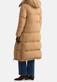 Lange, beige pufferjas met een capuchon en zichtbaar doorgestikt patroon, gecombineerd met blauwe wijde jeans en bruine haklaarzen.