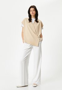 Top over-sized beige con design avvolgente, con dettaglio a bottone sul lato, abbinato a pantaloni bianchi a gamba larga e scarpe chiare.