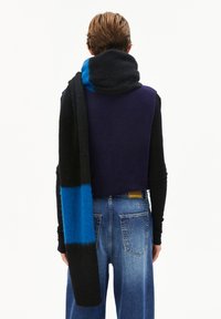 Personne portant un haut sans manches bleu foncé, une longue écharpe noire et bleu vif, des manches longues noires et un jean bleu ample, vue de dos.