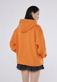 Pomarańczowy, oversize'owy hoodie z kapturem, charakteryzujący się luźnymi rękawami, miękką teksturą i swobodnym krojem. Noszony z czarną spódnicą z postrzępionym dołem.
