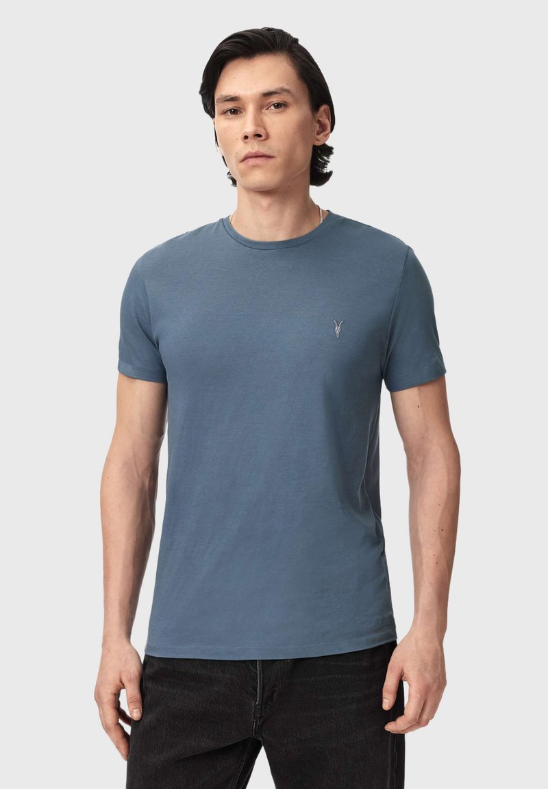 Homme portant un t-shirt bleu uni à manches courtes avec un petit logo sur le côté gauche de la poitrine, associé à un jean noir, devant un fond gris clair.