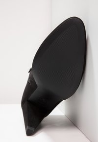 Musta mokkasiini korkokengät, joissa on terävä kärki ja teksturoitu pohja. Varusteena kapea kolmionmuotoinen korko ja minimalistinen muotoilu.