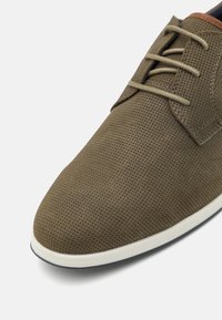 Chaussure en cuir perforé marron avec un bout arrondi lisse, semelle blanche contrastante et lacets beiges, présentant de subtils détails de couture.