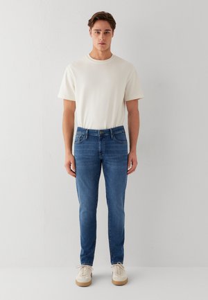 Junger Mann steht mit Blick nach vorne, trägt ein weißes kurzärmeliges Hemd, blaue Jeans und weiße Turnschuhe vor einem schlichten weißen Hintergrund.