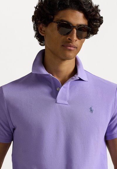 Μωβ πόλο μπλούζα με υφασμάτινη υφή, διαθέτοντας λαιμόκοψη με γιακά, πλαίσιο με δύο κουμπιά και ένα μικρό κεντημένο λογότυπο στο στήθος.