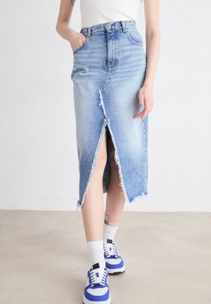 Femme portant une jupe en jean bleu clair avec une fente à l'avant et des bords effilochés, des chaussettes blanches, des baskets bleues et blanches, et un haut blanc.