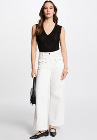 Top smanicato nero in maglia con scollatura a V, abbinato a pantaloni larghi bianchi a vita alta e ballerine nere. Gli accessori comprendono una borsa.