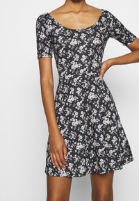 Robe noire à fleurs avec des manches courtes, un col rond et un corsage ajusté se terminant par une jupe évasée ornée de petites fleurs blanches et roses.