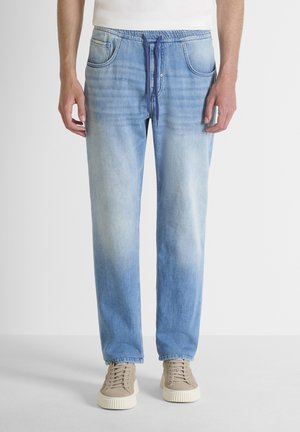 Man draagt lichtblauwe versleten denim joggerbroek met een marineblauwe trekkoord en beige sneakers tegen een witte achtergrond.