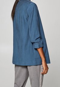 Blaue, leichte Jacke mit einem lockeren Schnitt, hochgekrempelten Ärmeln und schlichtem Design. Sichtbare Nahtdetails und Seitentaschen.