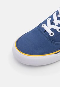 Gros plan sur le bout d'une basket en toile bleue avec des lacets blancs, une semelle en caoutchouc blanche et une fine bande jaune au-dessus de la semelle.