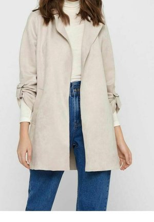 Vrouw die een beige openvallende jas met strikmouwen draagt, een witte coltrui en een blauw, hooggesloten spijkerbroek, staand tegen een effen achtergrond.