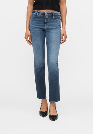 Femme portant un jean bleu droit, un top court noir sans manches et des talons noirs pointus, debout devant un fond uni.