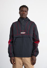 Cazadora negra con costuras y detalles en rojo, cuello con cremallera, bolsillo delantero y puños elásticos. Tejido ligero con un diseño deportivo.