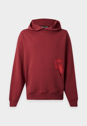 Hombre con sudadera roja con capucha, camisa de cuadros atada a la cintura, pantalones cortos negros, zapatillas negras, calcetines blancos, collar de cuentas, gorra y sosteniendo un bolso pequeño.