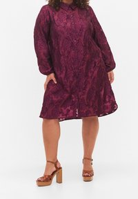 Burgundin väriinen mekko kukkakuviolla, pitkät puffihihaiset ja napit edessä; yhdistetty ruskeisiin platomainen sandaaleihin.