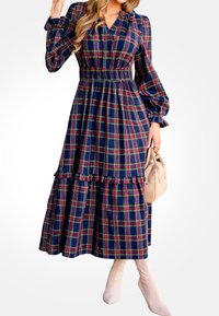 Largo vestido a cuadros en azul marino, verde y rojo, con una cintura fruncida, mangas abullonadas y un dobladillo con volantes. La modelo sostiene un bolso beige.