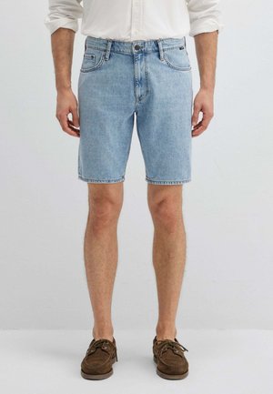 Mann trägt hellblaue Jeans-Shorts, weißes Langarmshirt mit hochgekrempelten Ärmeln und braune Veloursleder-Schnürschuhe, steht aufrecht.