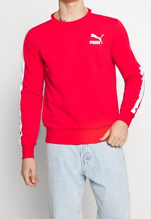 Mężczyzna w czerwonej bluzie Puma z białymi paskami na rękawach i jasnoniebieskich dżinsach, stojący na tle jednolitego tła.
