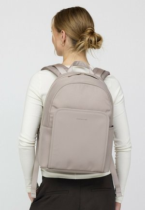 Frau mit blondem Haar zu einem Dutt gebunden, trägt ein weißes langärmeliges Oberteil und schwarze Hosen, trägt einen hellbeigen Rucksack mit mehreren Fächern.