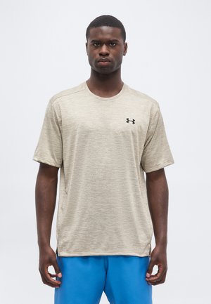 Jeune homme aux cheveux courts portant un t-shirt beige Under Armour à manches courtes et un short bleu, debout devant un fond clair uni.
