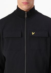 Chaqueta negra con cremallera y cuello alto, dos bolsillos en el pecho y un emblema dorado de águila en el bolsillo izquierdo. Textura de tela suave, diseño minimalista.