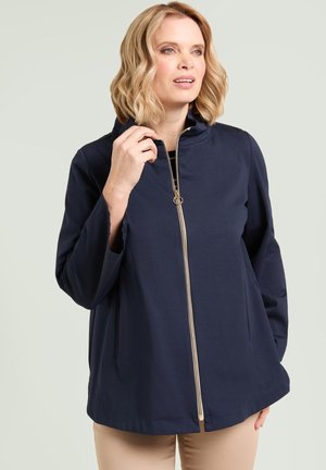 Blonde vrouw draagt een marineblauw jack met gouden rits, houdt de kraag vast, kijkt iets opzij, beige broek zichtbaar.