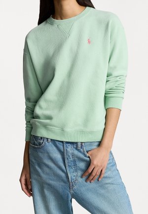 Kobieta w pastelowo zielonej bluzie z okrągłym dekoltem i różowym logo oraz jasnoblękitnych dżinsach, ręka spoczywa w kieszeni.