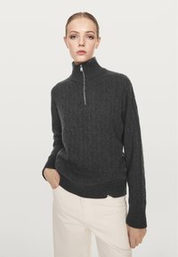 Vrouw met een donkergrijze kabelgebreide trui met een half-rits kraag en witte broek, staand met één hand in haar zak.