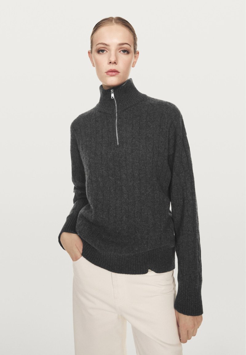 Vrouw met een donkergrijze kabelgebreide trui met een half-rits kraag en witte broek, staand met één hand in haar zak.