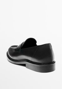 Mocassino in pelle nera con tacco basso, cucitura a moc toe e una fascia sopra il piede, mostrato da un'angolazione laterale posteriore.
