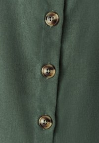 Oliivroheline kangas, millel on kolm pruunikat nuppu, igaühel kaks auku, taustaks tekstuuriga linane sarnane materjal.